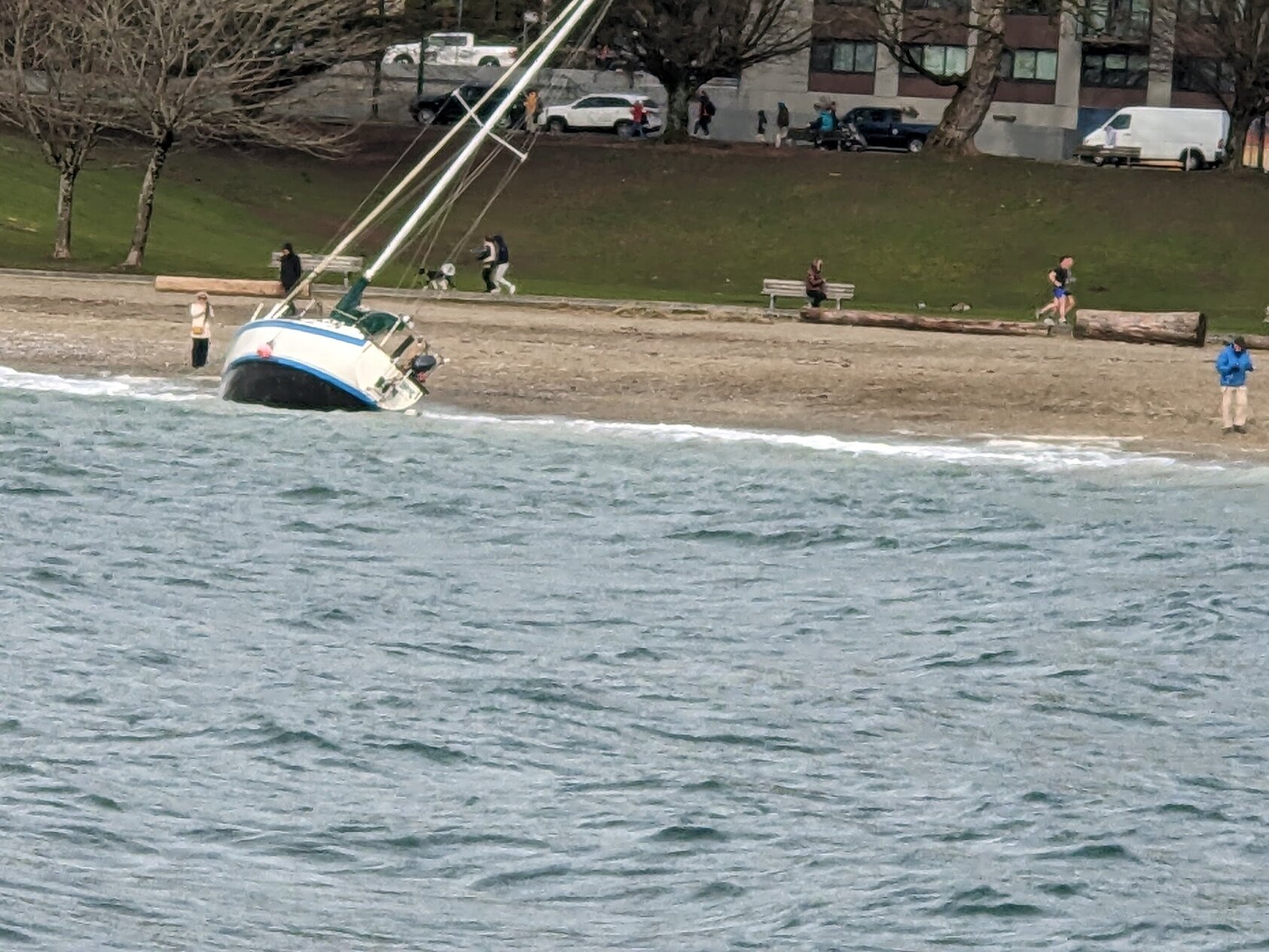 A white sailboat with blue trim lies at a 45° angle on a beach, its hull half-submerged in the waves.